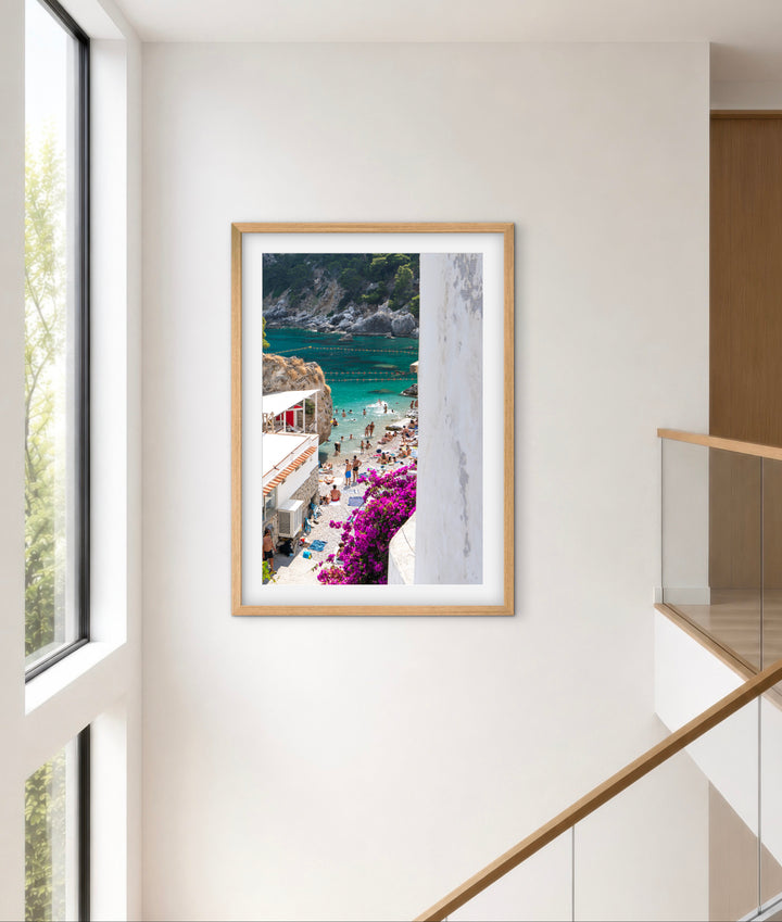 A framed art print depicting a scenic coastal village with turquoise water, white buildings, and bright purple flowers, displayed on a neutral wall in a modern interior setting.